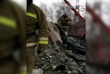 Two men in firefighter uniforms climb over a stack of rubble under a grey sky.