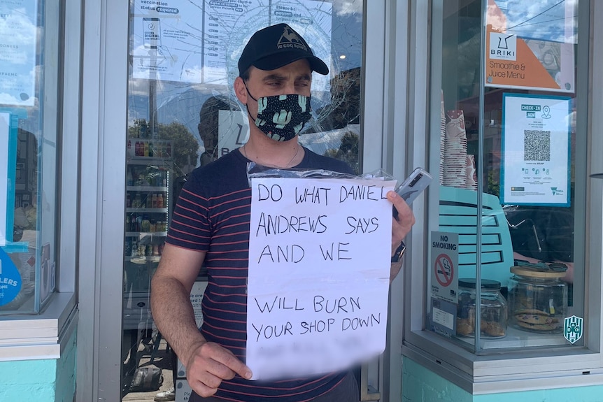 A man holds a sign that says "do what Daniel Andrews says and we will burn your shop down"