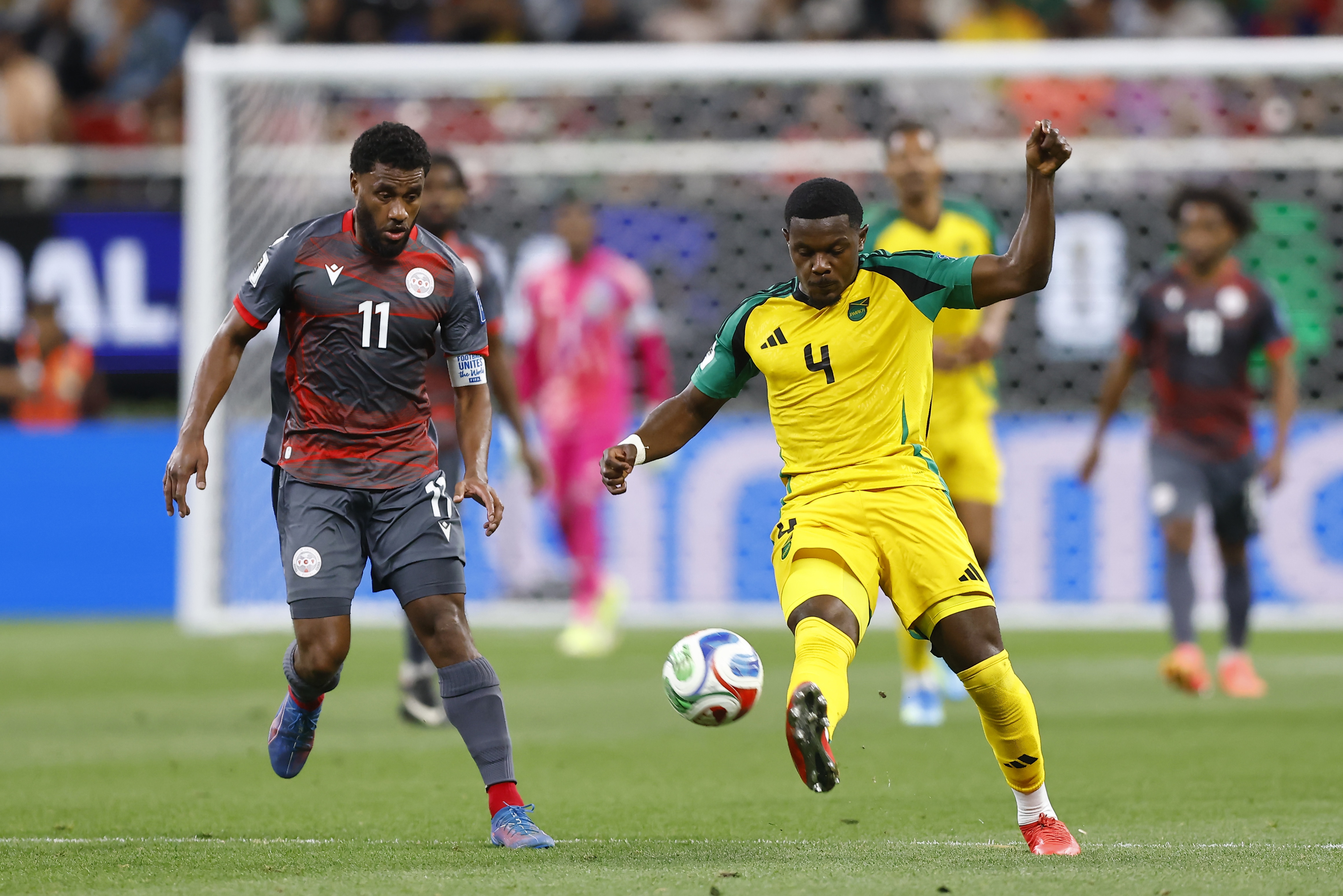 Um membro da seleção jamaicana de futebol masculino chuta a bola enquanto um zagueiro a persegue