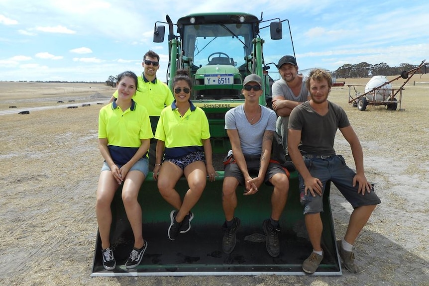 Six young farm workers sit in front of a tractor on a rural property.