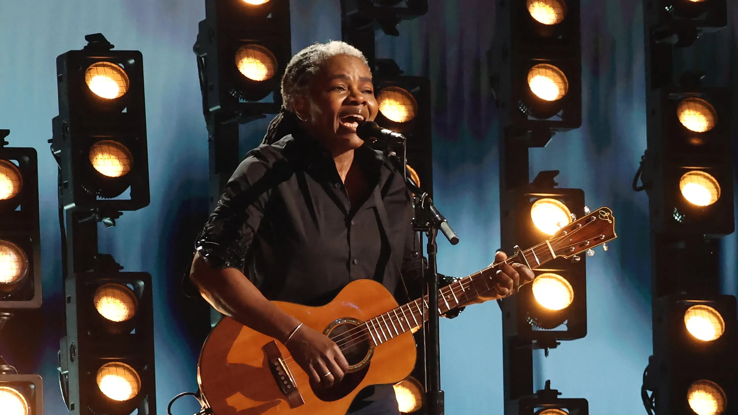 Tracy Chapman wears dark clothing and holds a guitar in front of backlights