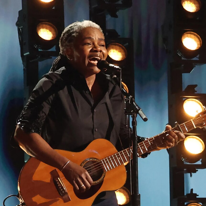 Tracy Chapman wears dark clothing and holds a guitar in front of backlights