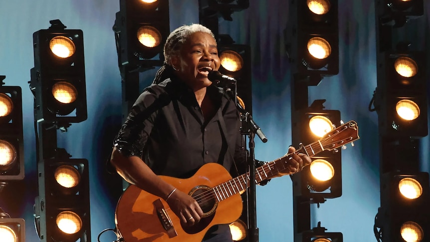Tracy Chapman wears dark clothing and holds a guitar in front of backlights