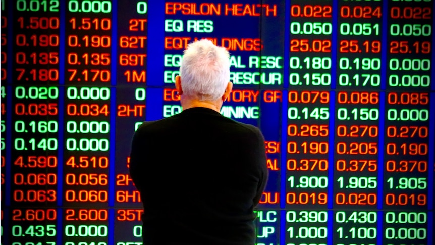 A man with white hair, and a black jumper, looking at the ASX stock market boards in Sydney.