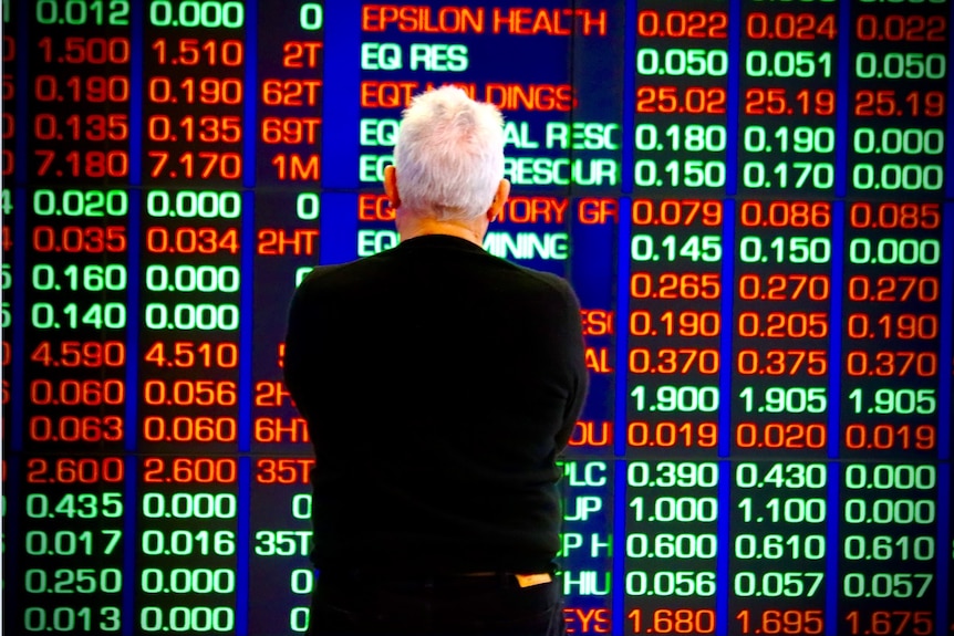 A man with white hair, and a black jumper, looking at the ASX stock market boards in Sydney.