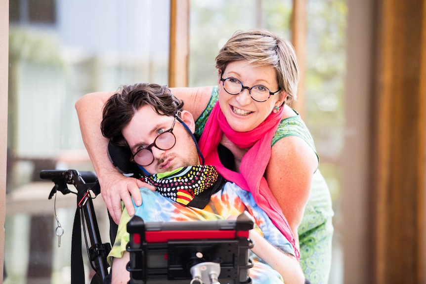 Jacob Hughes sits in a wheelchair. His mother stands behind him with her arms around his shoulders.