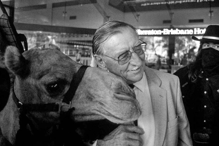 Black and white photo of smiling man in suit holding his face close to the face of a camel.