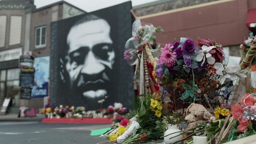 flowers on a street in the foreground with a black and white painting of george floyd in the background