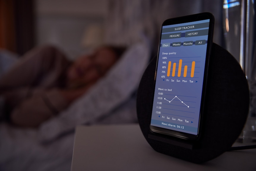 A photo of a phone on a bedside table with a sleep tracking app open. A woman is sleeping in the background