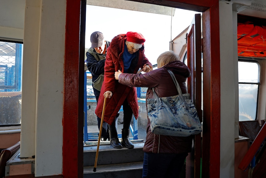 An elderley woman in a long burgunday coat is helped down stairs onto a ferry