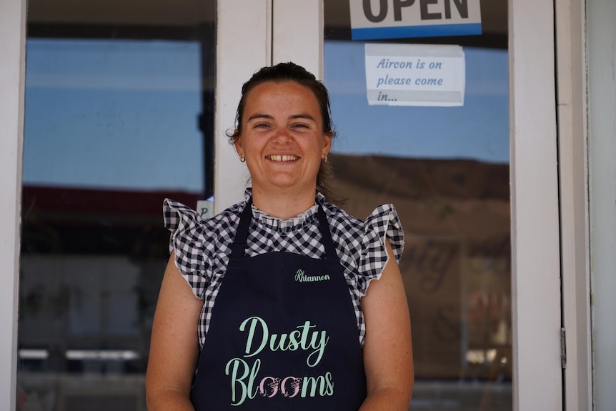 A woman with a blue apron stands in front of a door with an open sign