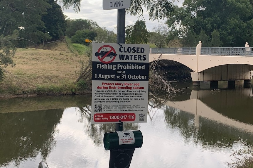 A sign, with a creek and a bridge in the background.