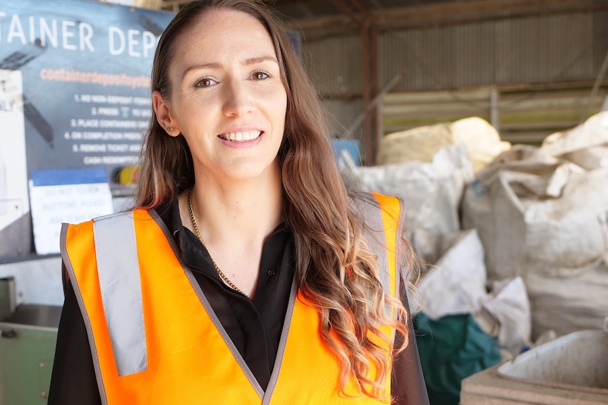 Tiffany Lim, wears orange high visibility vest, smiles, bags of containers in background inside depot.