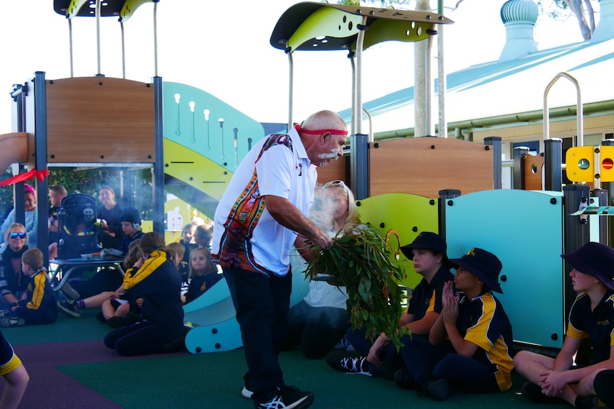 Aboriginal man with red headband holds a large eucalyptus branch smoking walking in front of children at the new playground