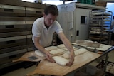 A male baker shapes dough into rectangle shapes on a bench inside a bakery.