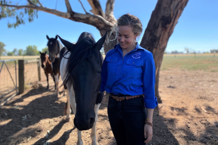 A woman in a blue work shirt smiles at a black and white horse standing next to her in a paddock. 