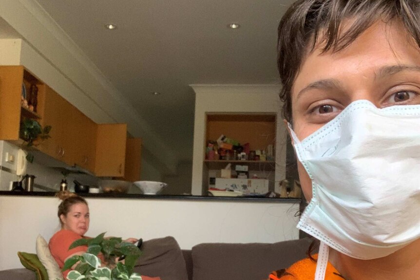A woman wearing a face mask in the living room of her home with another woman sat on a couch behind her
