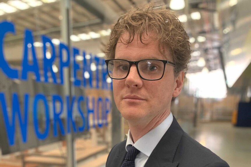 Peter Hurley, wearing glasses, a grey suit and blue tie, standing front of a window with 'carpentry workshop' on it