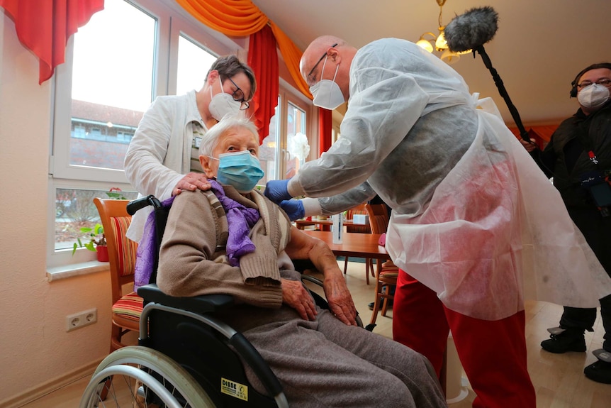 Two doctors in white gowns and face mask inject elderly white-haired man in wheelchair in room.