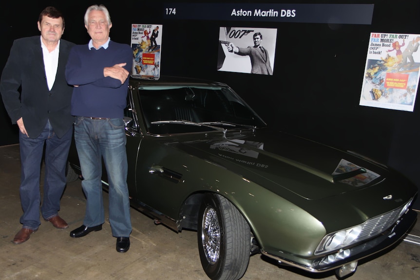 Two men stand next to an olive green sports car