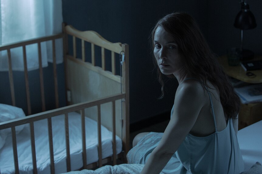 Woman sits nervously on the edge of a bed next to a cot, wearing a white silk nightie and bathed in cool blue light.