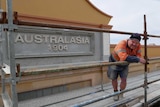 A man leans against structural beams with a concrete sign reading 'Australasia 1904' in the background.