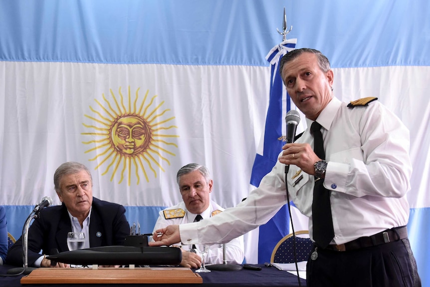 a man stands holding a microphone with two men sitting at a desk behind him with a flag behind them