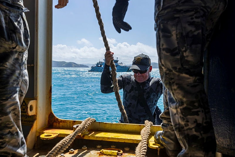 A man in a camo wet suit pulls himself up with a rope and into the open doorway of a a boat.