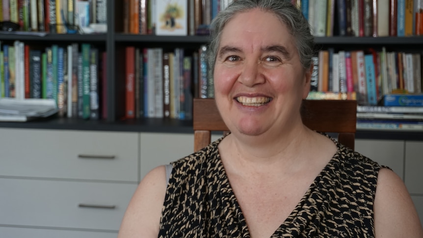 A smiling woman sits in front of a bookshelf.