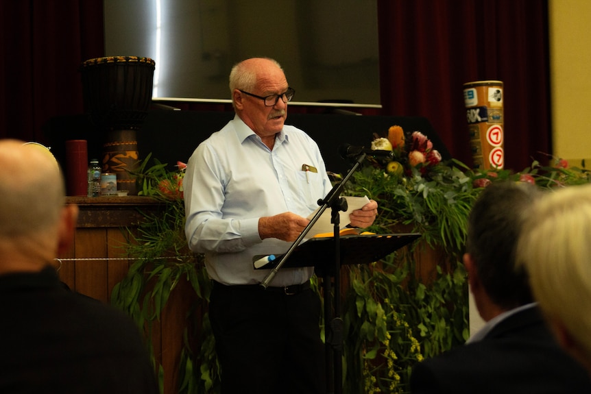 A man in his 70s wearing glasses reads from a piece of paper at a lectern with flowers and musical instruments behind him.