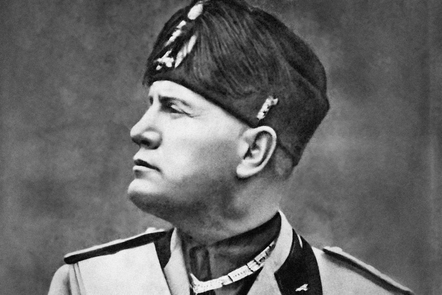 Black and white photo of Benito Mussolini, in uniform and head turned in profile, with large feather in hat.