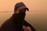 Gus Goswell wearing a hat and shirt over his nose sits near the water in Mallacoota.