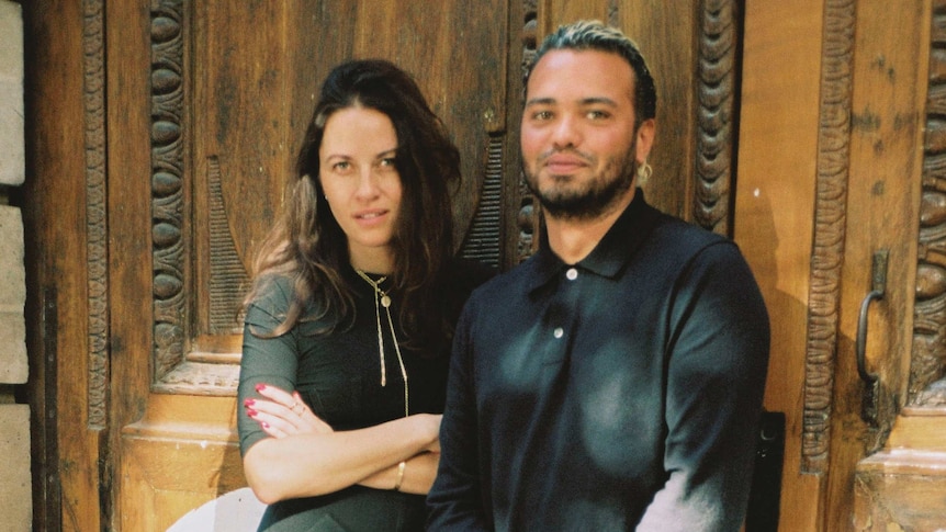 Kym Ellery and Duran Lantink stand looking in front of an ornate carved wooden door, smiling at the camera.