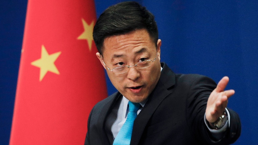 A man in a suit holds out his hand while standing in front of a Chinese flag