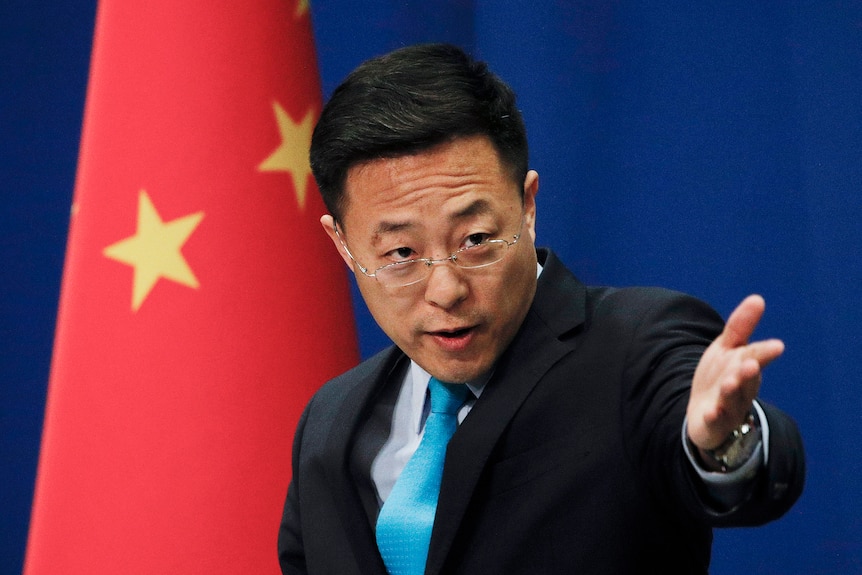 A man in a suit holds out his hand while standing in front of a Chinese flag