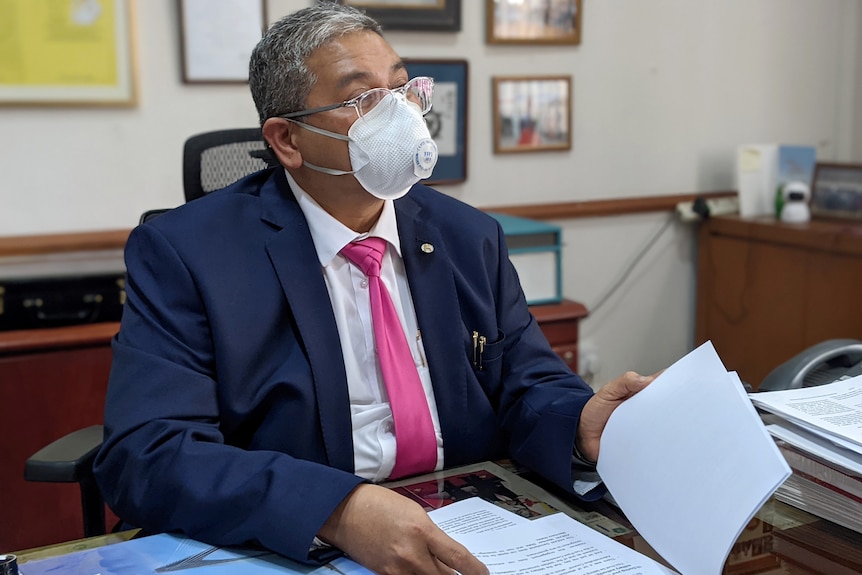An elderly southeast Asian man wearing a face mask in dark blue suit leafs through papers at his desk