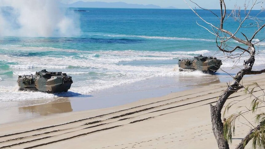 Amphibious military assault vehicles travel through the ocean to land on a beach.