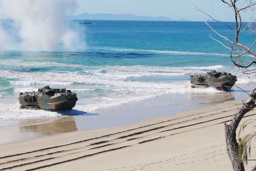 Amphibious military assault vehicles travel through the ocean to land on a beach.