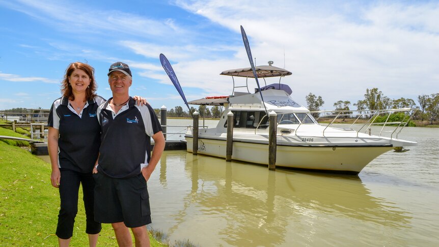 A man and a woman stand with each other smiling on a grassy bank next to a cruiser on the Murray River. 