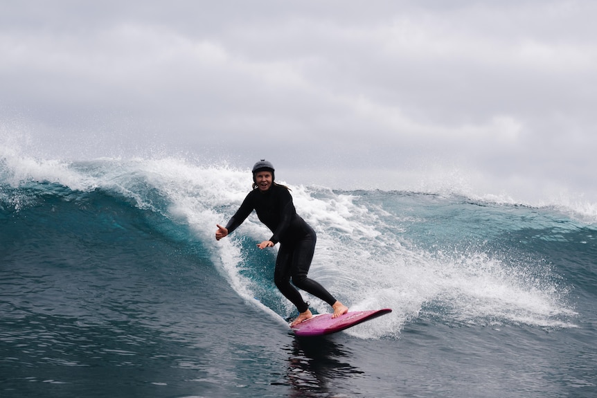 A woman wearing a black full length wetsuit is riding a wave, standing on a pink surfboard