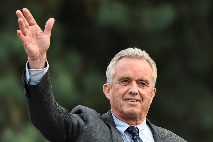 An older, cleanshaven white man in a grey suit waves with his right hand in front of some green trees.