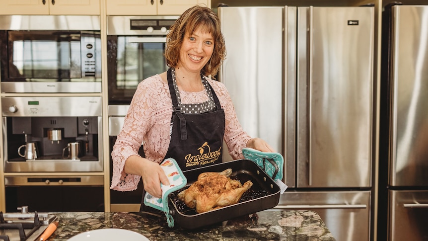 A woman wearing an apron is grinning standing in a kitchen holding a roast chicken in its baking tray 