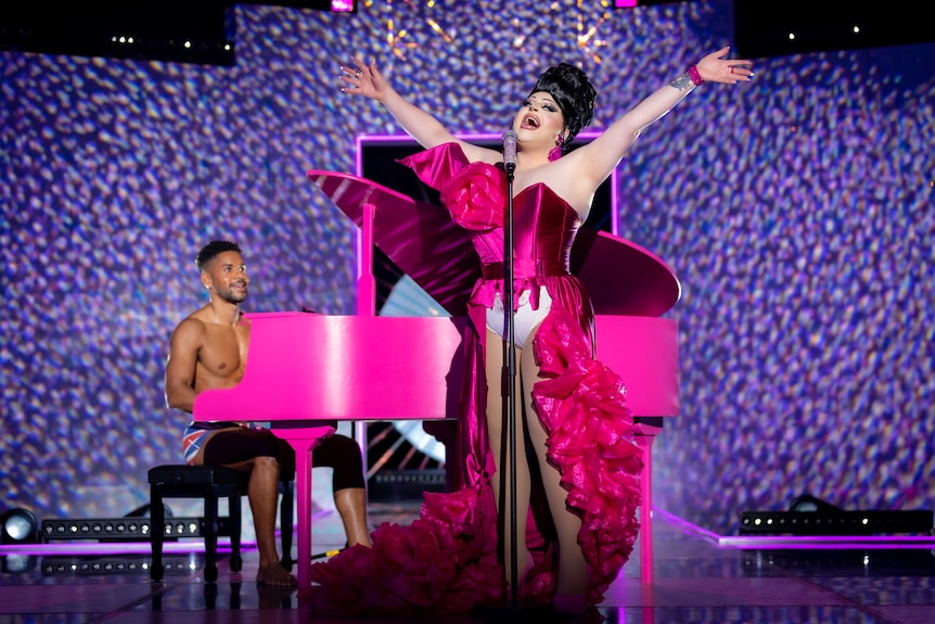 Hannah sings into a microphone in a frilly pink dress with white underwear on display. A shirtless man plays a pink piano. 