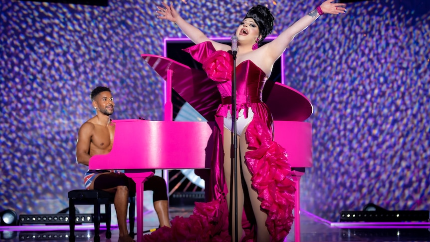 Hannah sings into a microphone in a frilly pink dress with white underwear on display. A shirtless man plays a pink piano. 