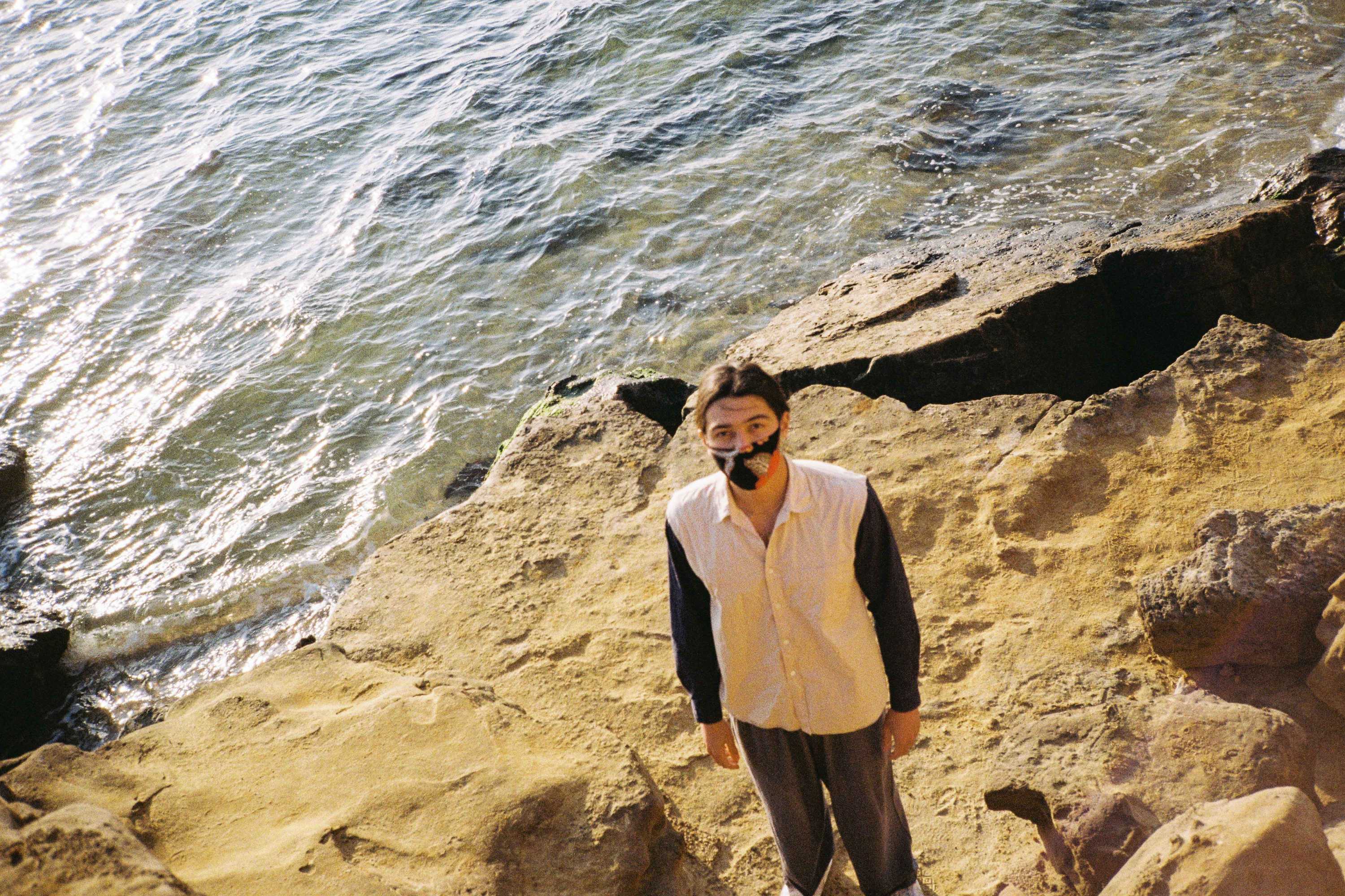 a photo of kita standing on a rocky edge next to the sea, the photo is taken from above and he's lookingup at the camera