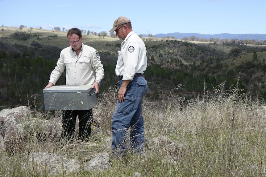 Two men in khaki ranger uniforms stand in grass in front of a hill. One man holds a large steel box.