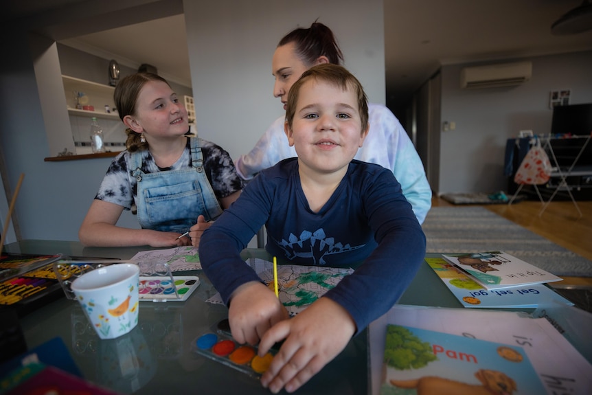 A young boy sits at a table smiling with his sister and mum behind