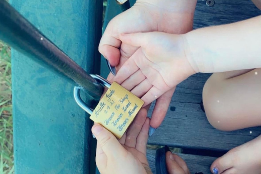 A padlock with inscription attached to a railing with a woman’s hands and two child hands holding it