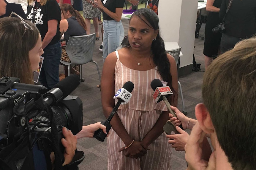 A woman speaks to reporters, a number of people gather in the background.