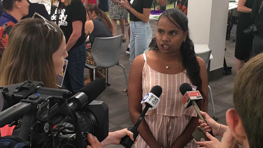 A woman speaks to reporters, a number of people gather in the background.